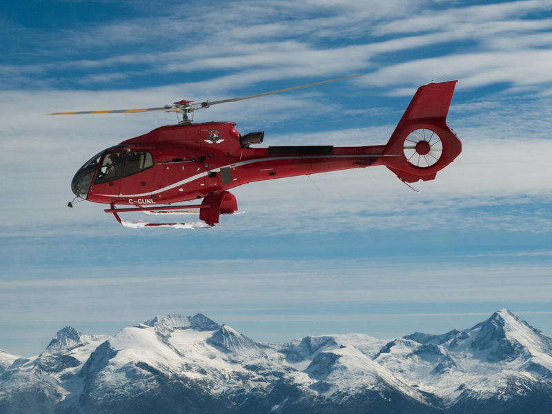 Red helicopter flying above the snow-covered Coast Mountains on a Whistler Blackcomb helicopter tour under a bright blue sky.