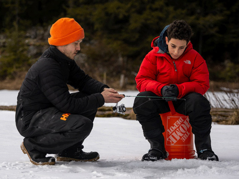 A guide in an orange toque shows a young angler in a red jacket how to set up an ice fishing rod on a frozen lake near Whistler.