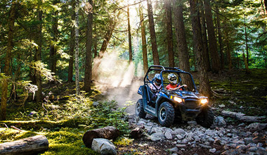 Side-by-side ATV drives over a rocky forest trail in Whistler, kicking up dust through sunlit trees.