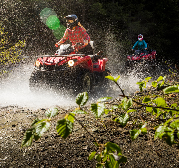 Two people ride red ATVs through a muddy forest trail in Whistler, splashing water as they speed through a puddle, surrounded by dense trees and sunlit spray.