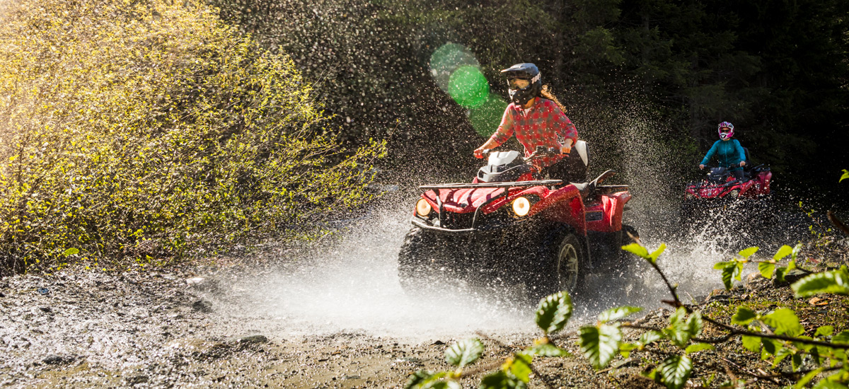 Two people ride red ATVs through a muddy forest trail in Whistler, splashing water as they speed through a puddle, surrounded by dense trees and sunlit spray.