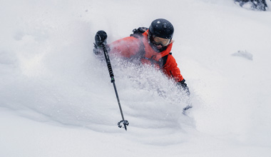 Close-up of a skier in a bright red jacket and black helmet charging through deep powder, face partially covered by swirling snow as they carve a dynamic turn down a steep slope in Whistler.