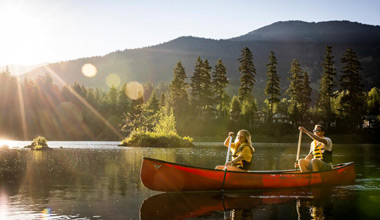 Two people paddle a bright red canoe across a calm lake at golden hour, sunlight flaring over the Coast Mountains and reflecting off the glassy water as evergreen trees line the shore.