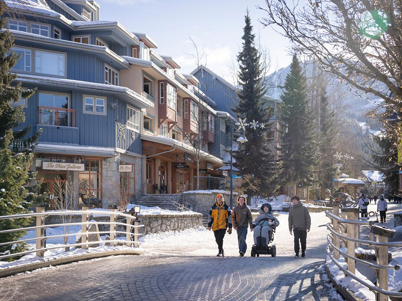 A group of friends, including one in a wheelchair, enjoy a sunny winter day in Whistler Village, with snow-covered mountains and cozy accommodations in the background.