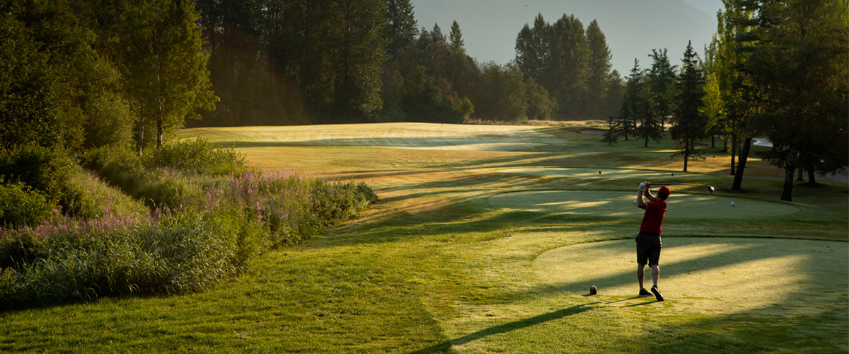 A golfer tees off on a sunlit fairway in Whistler, surrounded by tall evergreen trees and soft morning light casting long shadows across the manicured course.