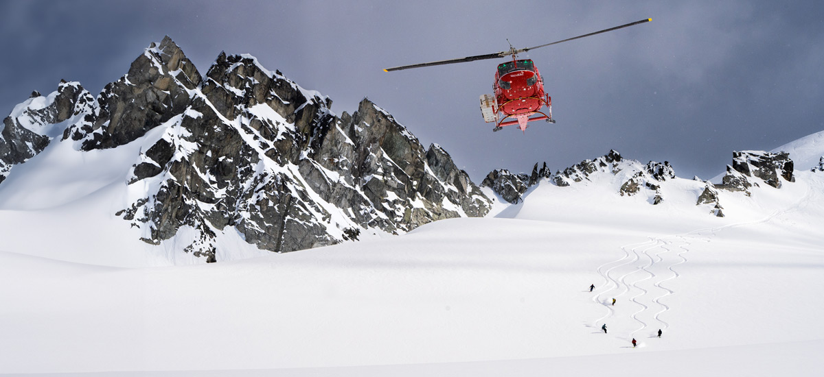 Heli-skiers watch a red helicopter approach above a snowy alpine ridge in Whistler’s backcountry, surrounded by rugged Coast Mountains and drifting snow.