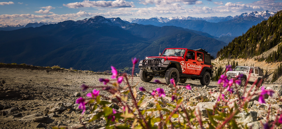 Off-road Jeep tour on a rocky alpine trail high above Whistler, with a red 4x4 in the foreground and a second vehicle behind, framed by pink wildflowers and sweeping Coast Mountain views under a dramatic sky.
