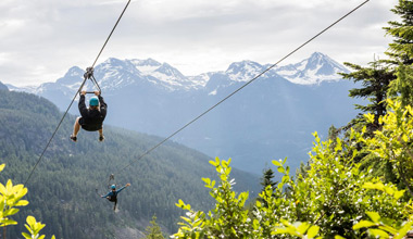 A rider drives an ATV along a gravel mountain road in Whistler, kicking up dust in the warm evening light with alpine peaks silhouetted behind.