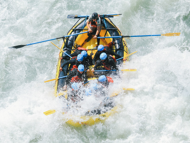 Five rafters paddle a yellow raft through churning whitewater on a tree-lined river, smiling and working together as waves splash around them.