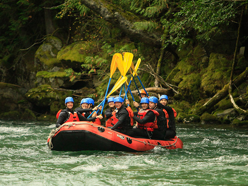 Group of rafters in blue helmets and red life jackets paddle a red inflatable raft through a green river, smiling and raising yellow paddles against a backdrop of moss-covered rocks and dense forest.