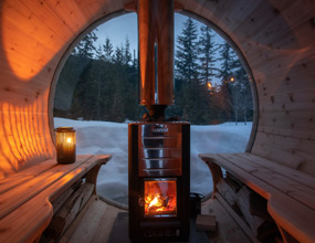 Wood-lined sauna interior with two curved benches facing a black wood-burning stove, its fire glowing warmly beneath a tall chimney. A large circular window frames snowy trees and a mountain landscape at dusk, with a lantern casting soft light inside.