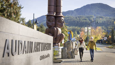 Two visitors walking past the Audain Art Museum sign and carved totem pole sculpture in Whistler Village, with rugged mountain peaks in the background.