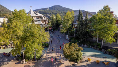 Sunny summer day on Whistler’s Village Stroll, with people walking among shops, trees, and mountain views in the background.