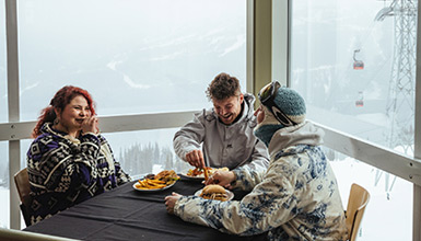 Three people dining at the Roundhouse Lodge on Whistler Mountain with the Peak 2 Peak Gondola in the background