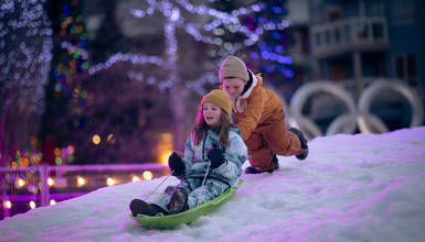 Two children sledding on snow under twinkling village lights, one steering while the other pushes with joy — a playful winter evening in Whistler Village.