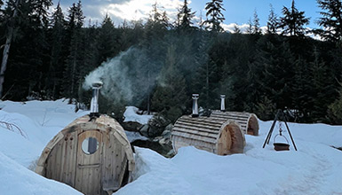 Three rustic, wood-fired barrel saunas sit in a snowy alpine setting in Whistler, British Columbia. Steam rises into the crisp mountain air, surrounded by evergreen trees and a serene winter landscape. A metal tripod with a hanging pot adds to the cozy, off-grid atmosphere.