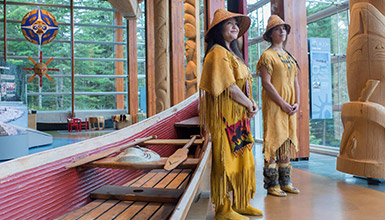 Two Indigenous cultural ambassadors wearing traditional cedar-bark regalia standing beside a handcrafted canoe and carved totem poles inside Whistler’s Squamish Lil’wat Cultural Centre.