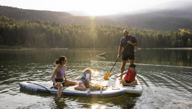 Family paddleboarding on Whistler's Lost Lake at sunset, surrounded by forested mountains and golden summer light.