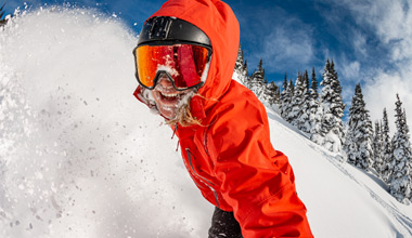 Close-up of a skier in a bright red jacket and black helmet charging through deep powder, face partially covered by swirling snow as they carve a dynamic turn down a steep slope in Whistler.