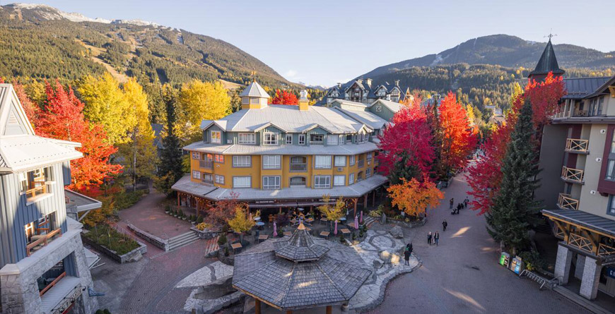 An aerial view of Whistler Town Plaza on a sunny fall day with red, yellow and orange foliage on the trees and people walking on the stroll.