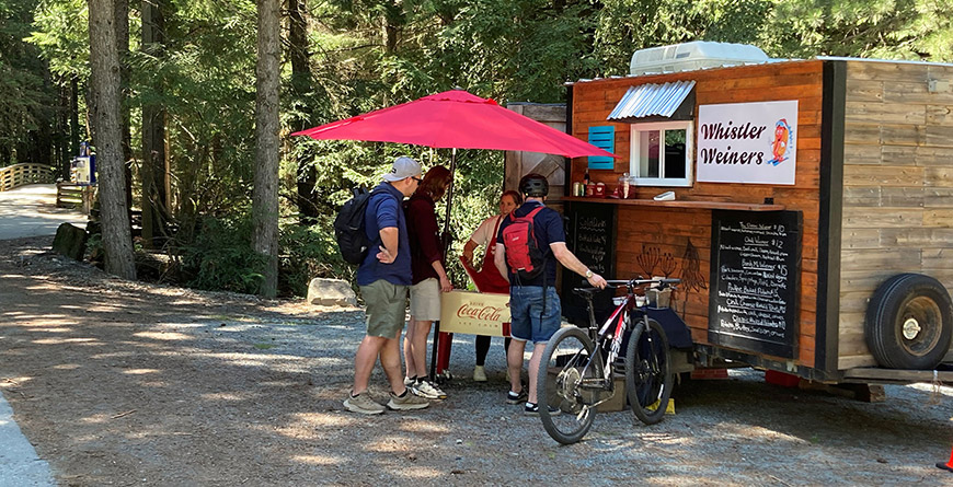 Group ordering from the Whistler Weiners food truck along a forested trail, with a bike parked nearby and a red umbrella providing shade.