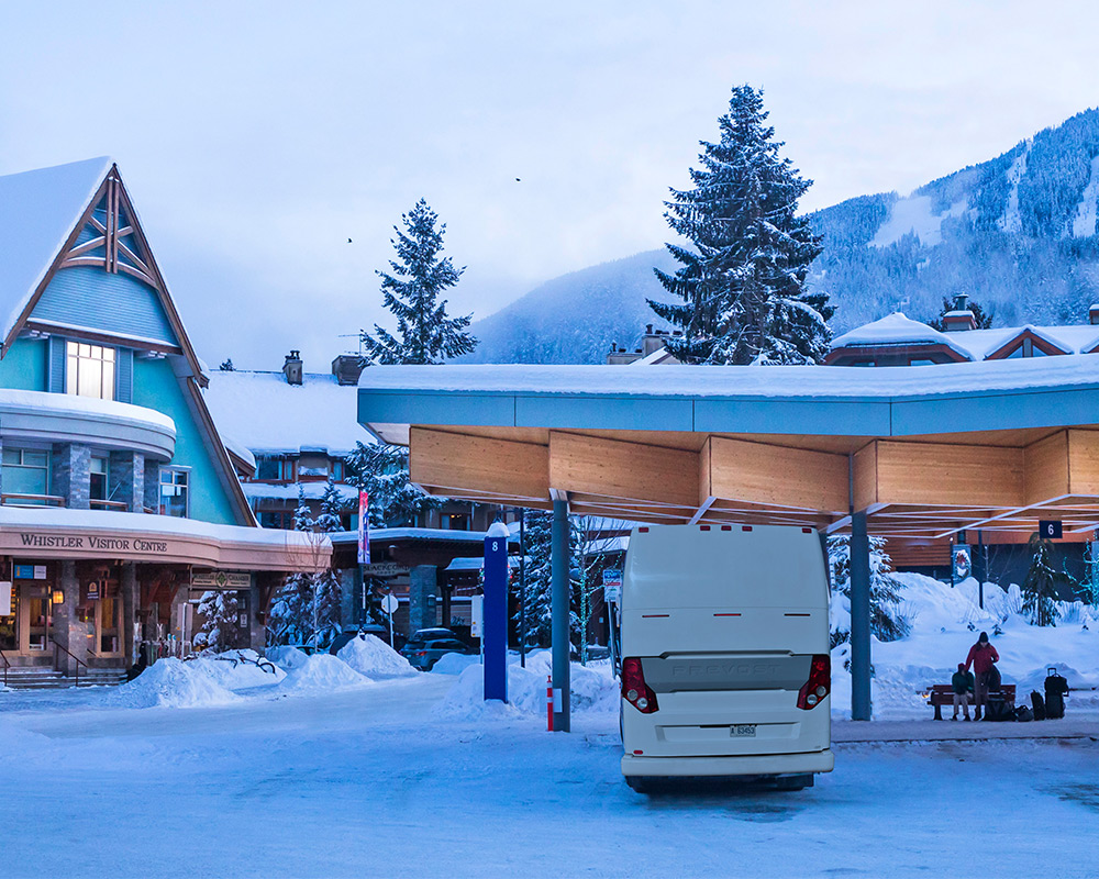Shuttle bus at snowy Whistler transit station with mountain and pine trees in background.