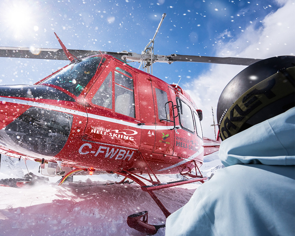 Skier watches red helicopter land in snowy Whistler backcountry.