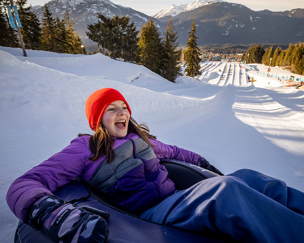 Smiling child snow tubing down sunny slope at Whistler’s Tube Park.